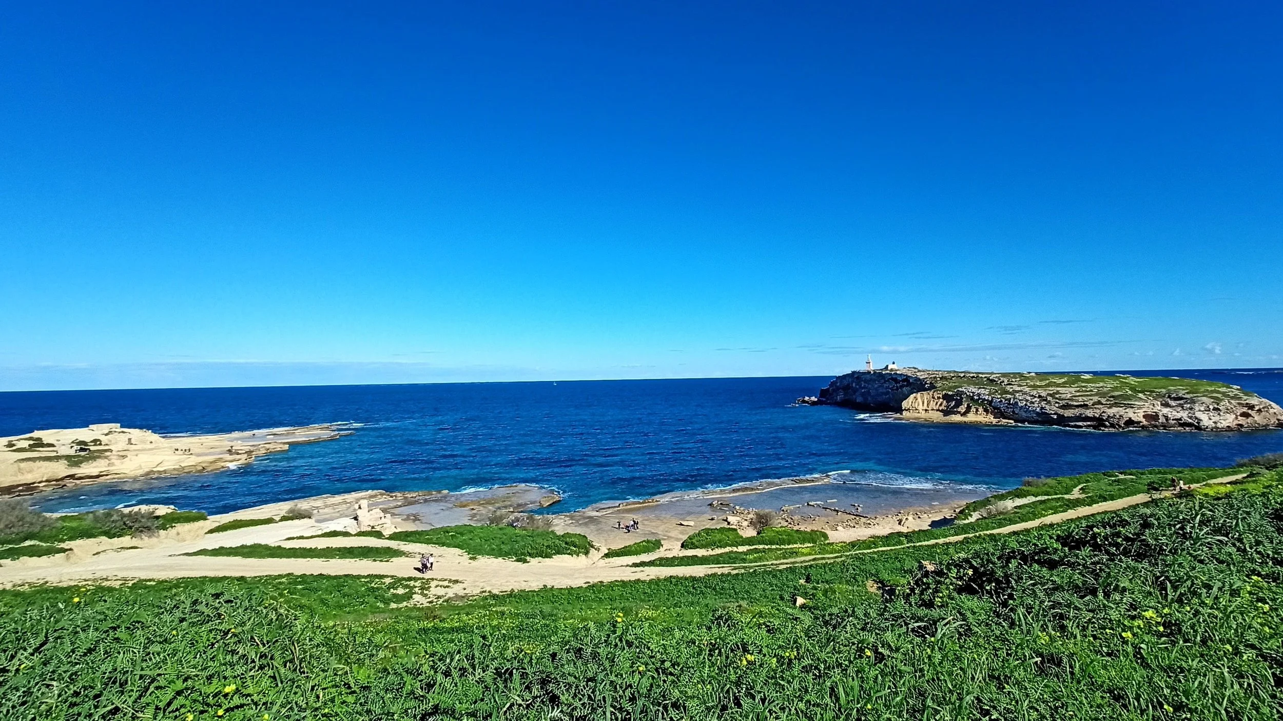 Scenic coastal landscape with green grassy hills, sandy paths, rocky beaches, and blue ocean waters under a clear blue sky. There are small figures of people on the beach and a lighthouse on a rocky promontory in the distance.