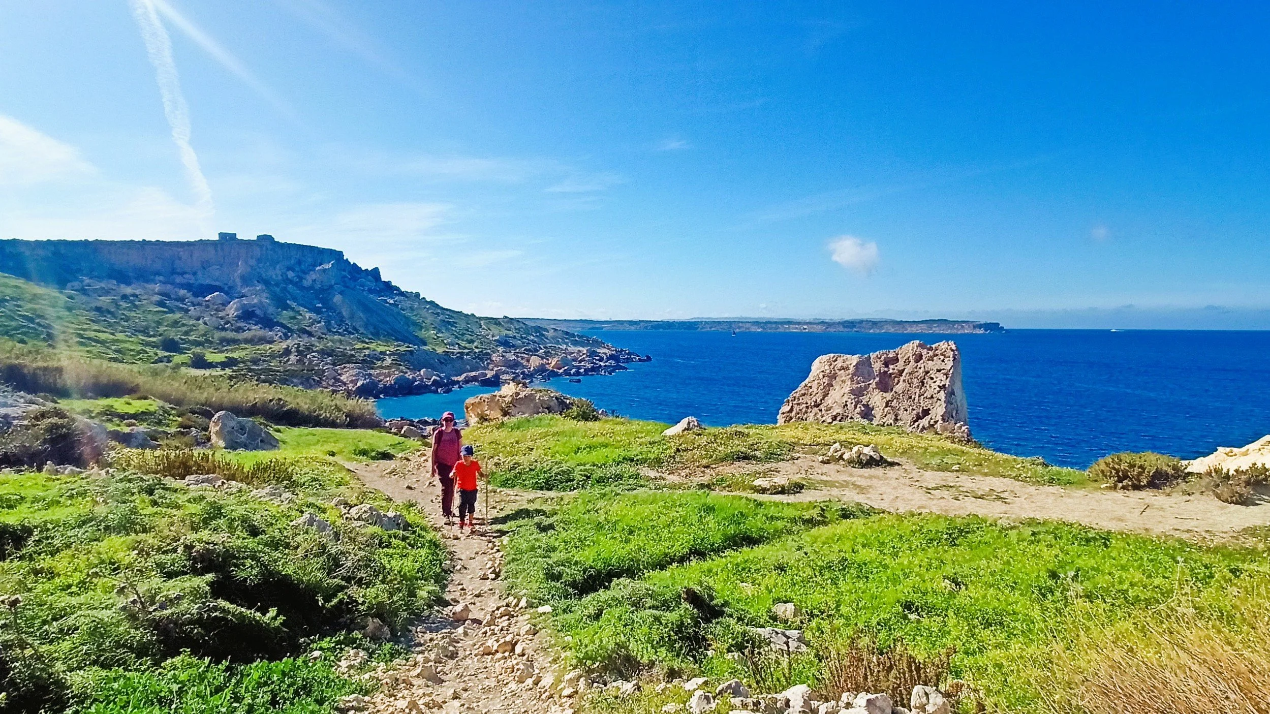 A woman and a child walking along a dirt trail on a green hillside overlooking a blue ocean with a rocky coastline and cliffs in the distance under a clear blue sky.