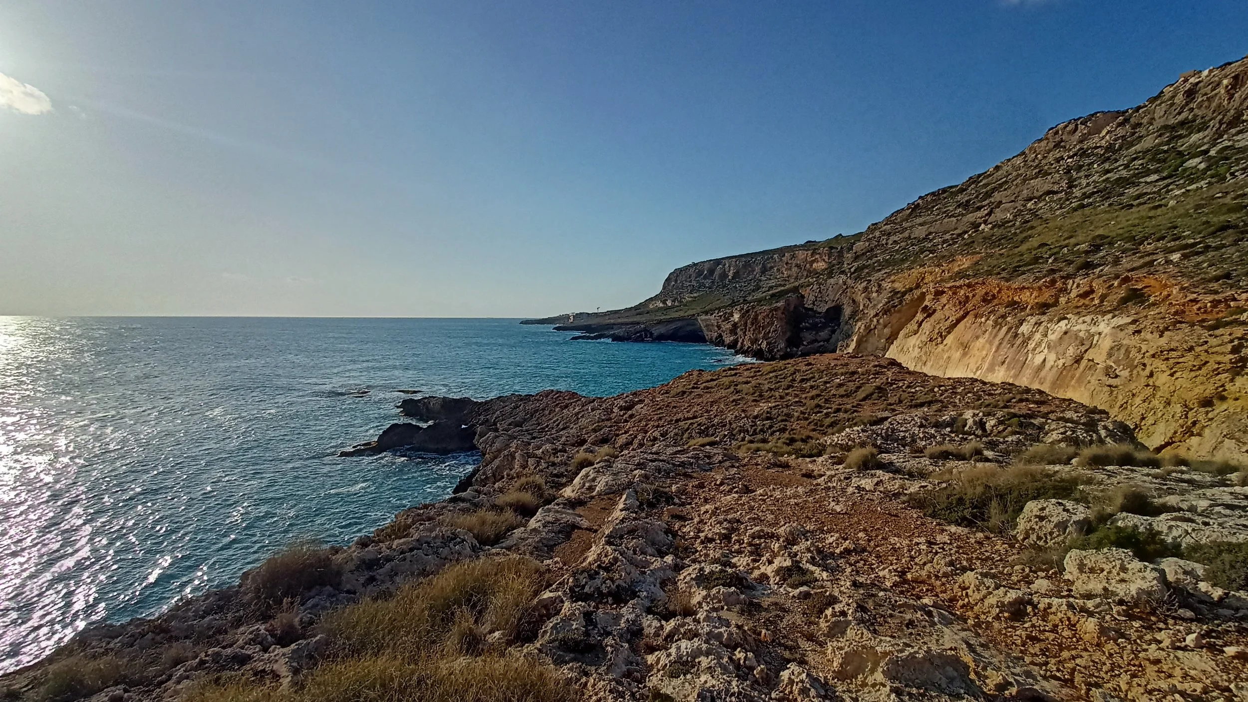 Coastal landscape with rocky shore, cliffs on the right side, and the ocean on the left, under a clear blue sky with the sun shining.