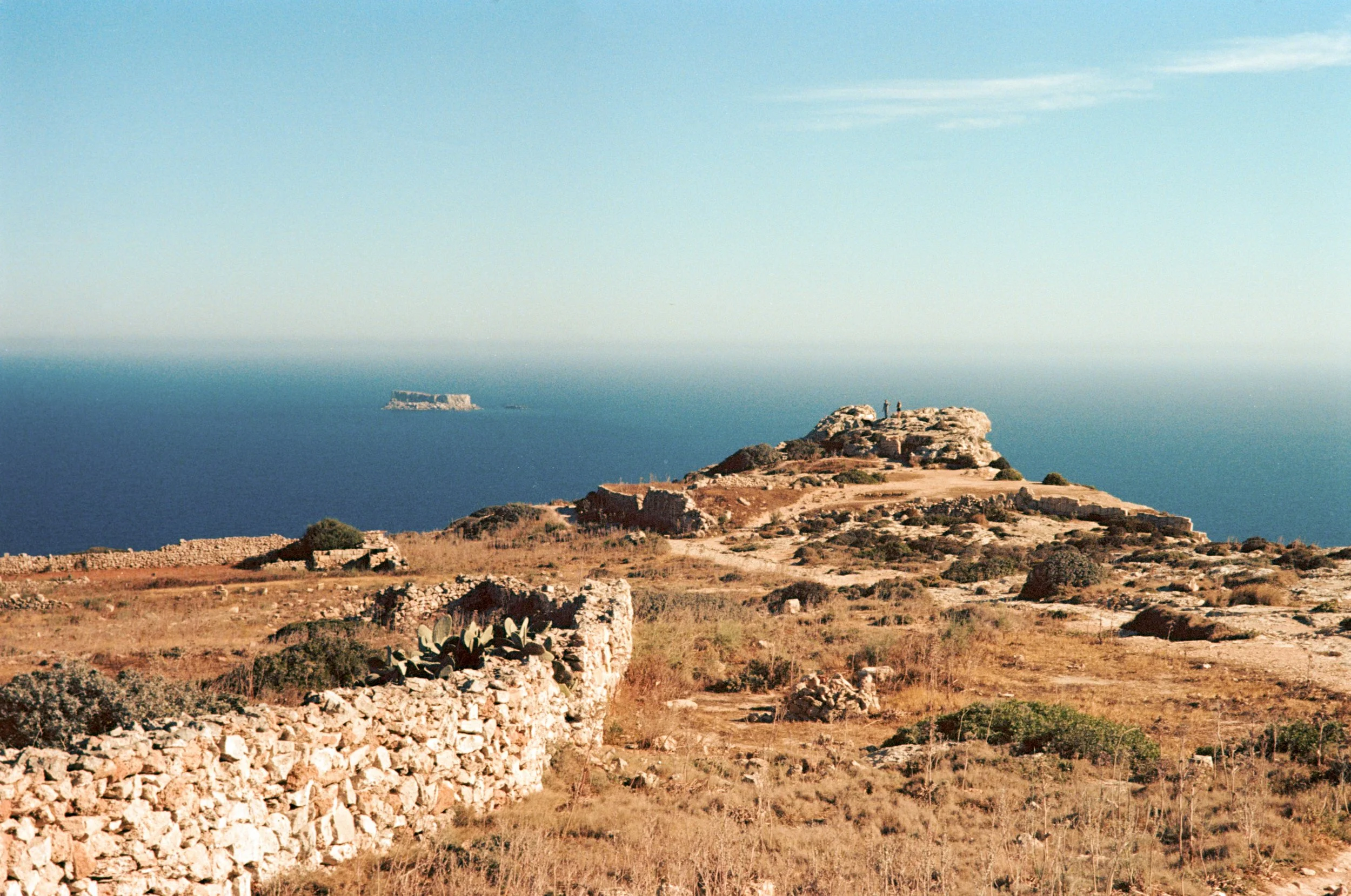 A landscape of a dry, rocky coastal area with ruins and cactus plants, overlooking the blue sea with a small island in the distance under a clear sky.