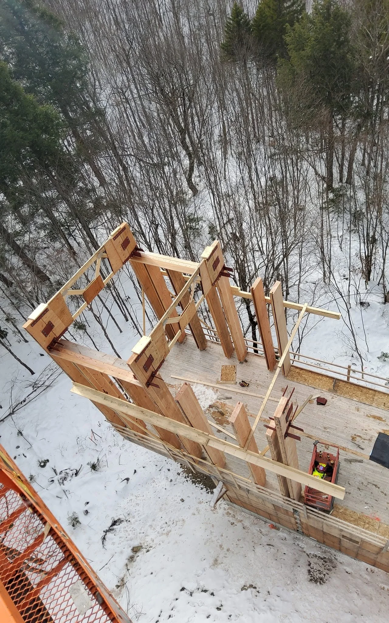Construction site of a wooden house frame surrounded by snow, with a forest of trees in the background.
