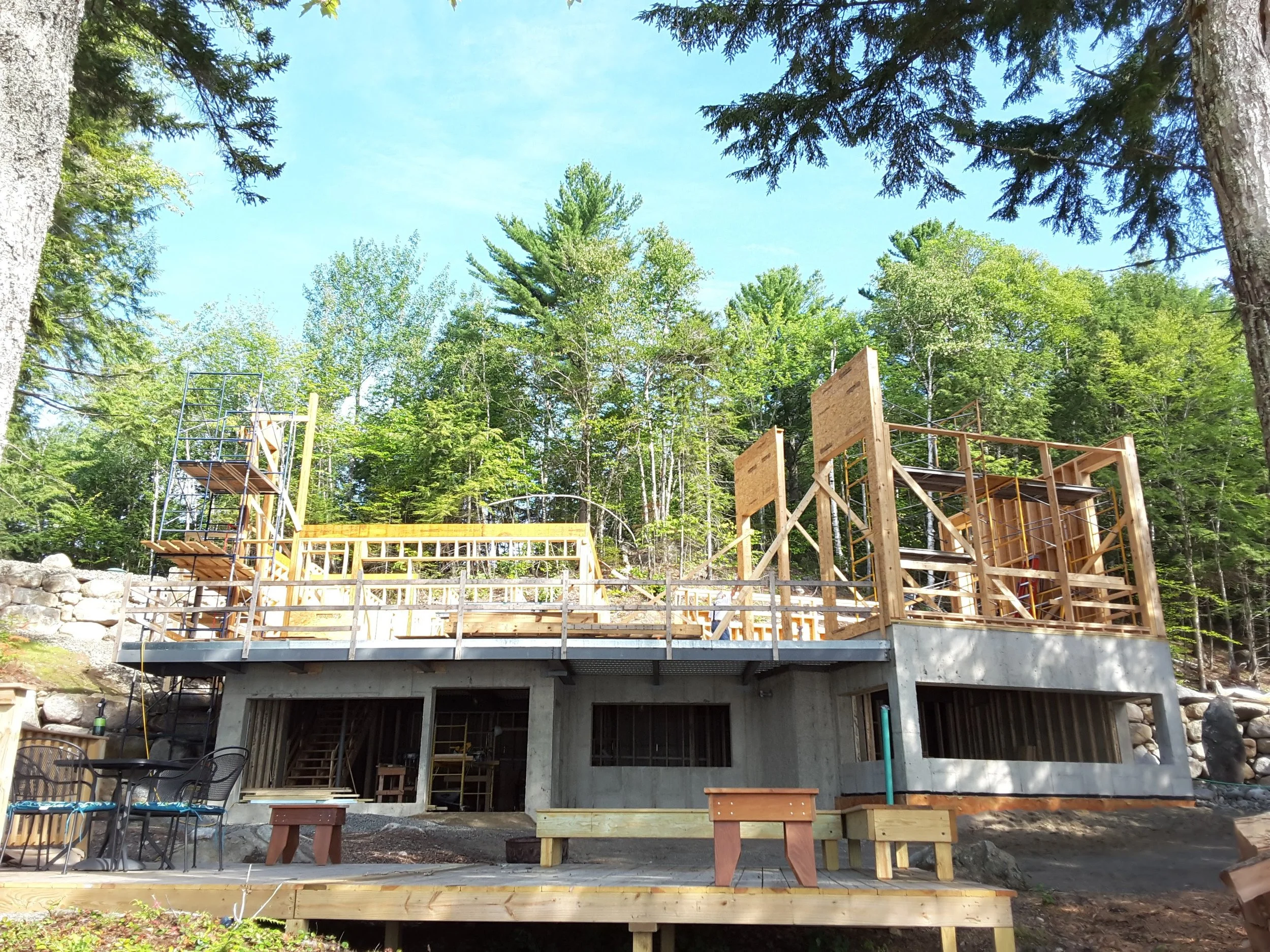 Construction site on a forested hillside with wooden framing and scaffolding, under a clear blue sky.