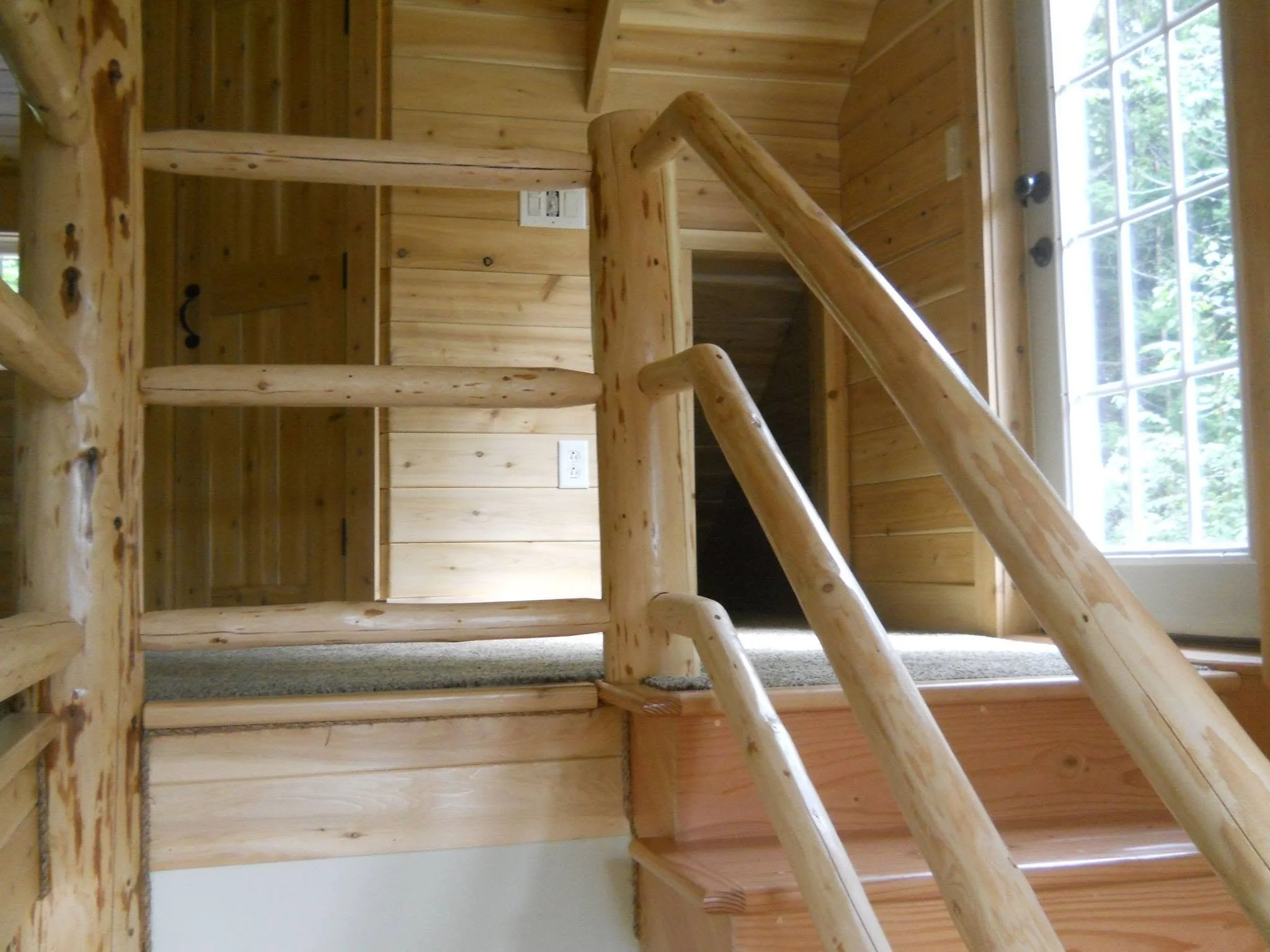 Interior view of a wooden staircase with a log railing, leading up to a small landing near a window with natural light.