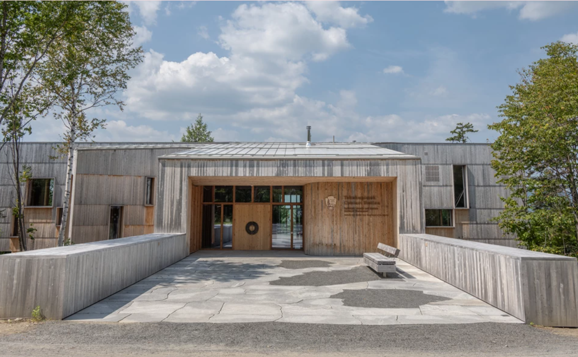 Modern building with wooden and metal exterior, surrounded by trees, featuring an entrance with large glass windows and a wooden door, on a paved area with a bench nearby.