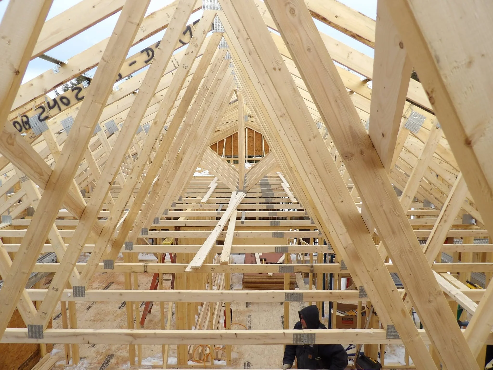 The image shows the wooden framework of a building under construction, with diagonal and horizontal beams forming the roof structure. There are metal brackets connecting the beams and a person working on the construction site.