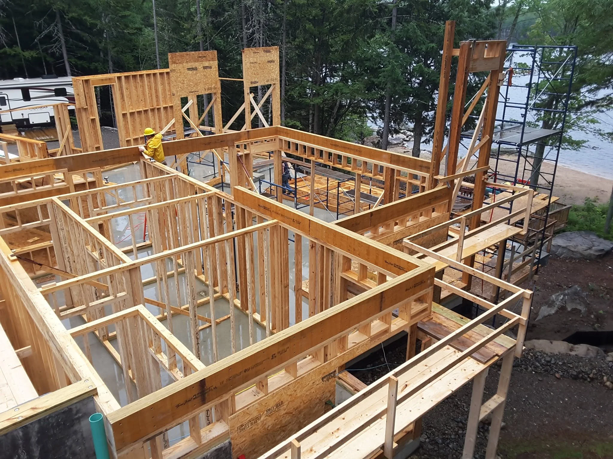 Construction framing of a house near a lake with trees in the background, showing wooden wall and floor structures, scaffolding, and a worker in a yellow jacket and helmet.