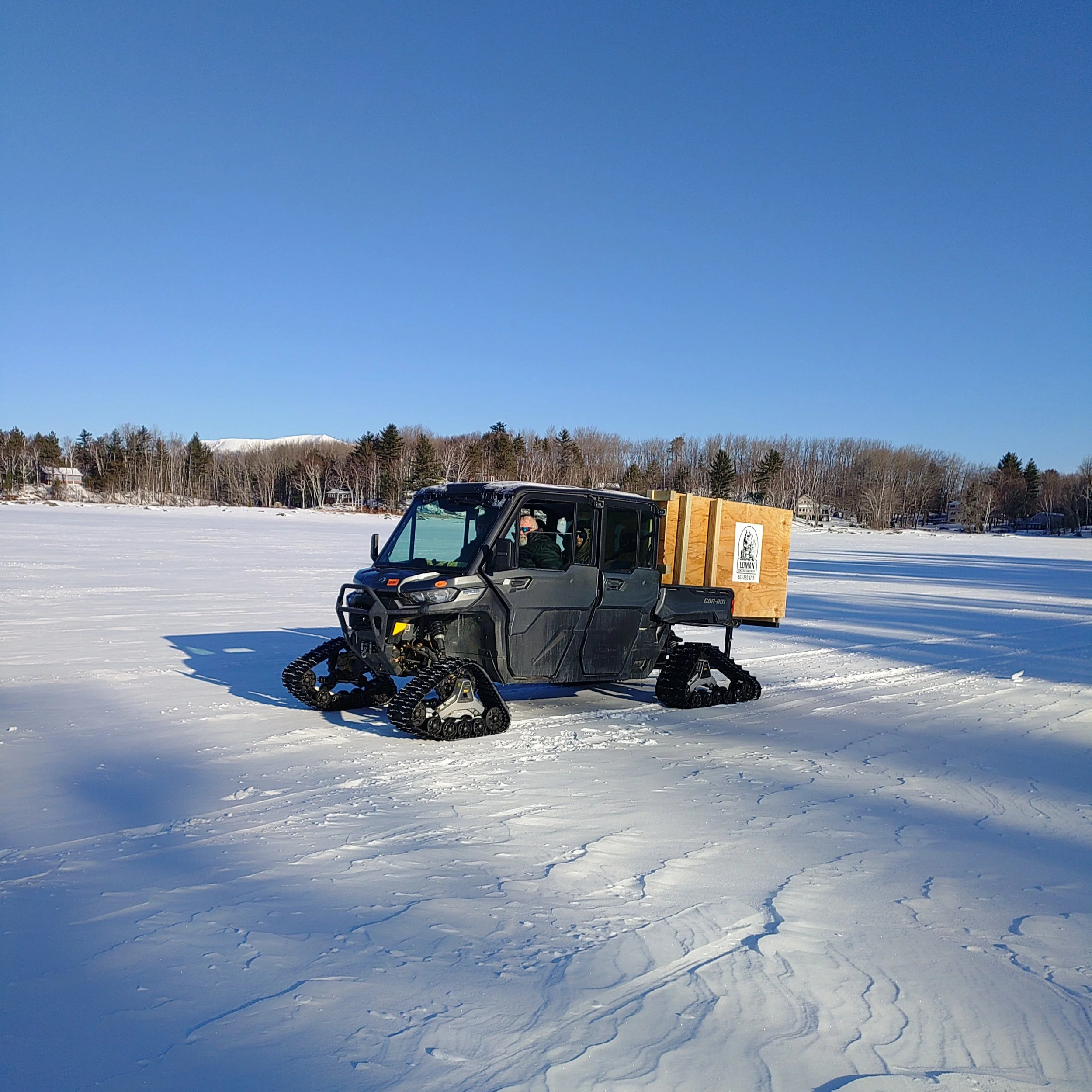 A snow-covered landscape with a large clear blue sky. In the foreground, a black snowmobile is visible with a wooden crate attached to its back, containing a logo and text that is partially readable. In the background, there are trees and some buildings visible at the horizon.