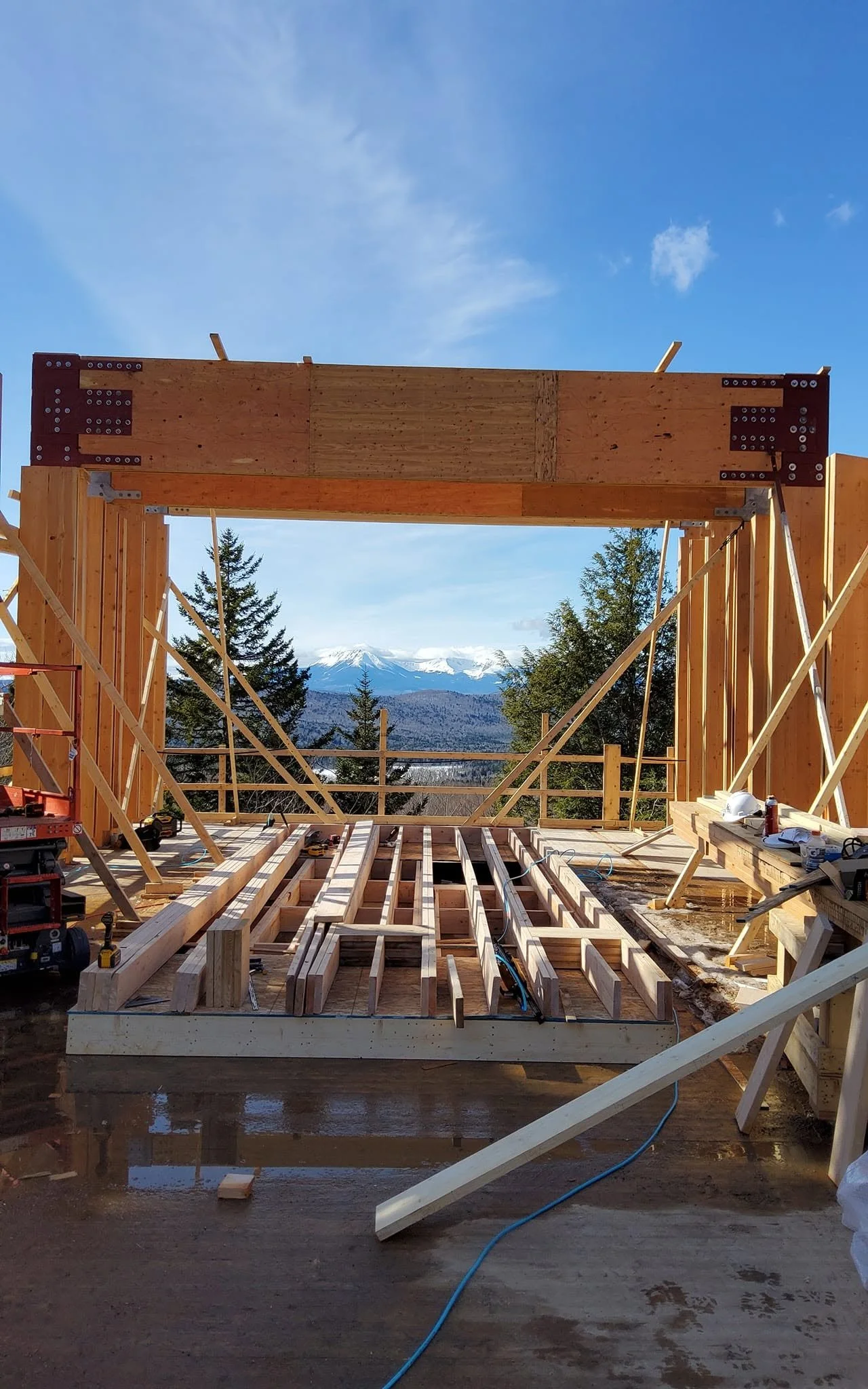 Construction site with wooden framing and beams, view of mountains and trees in the background under a clear blue sky.