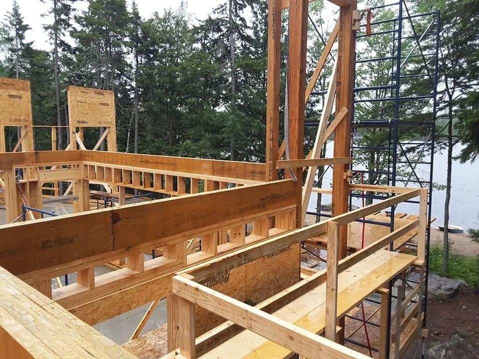 Construction site of a wooden house near a lake, with scaffolding and framework in progress, surrounded by trees.