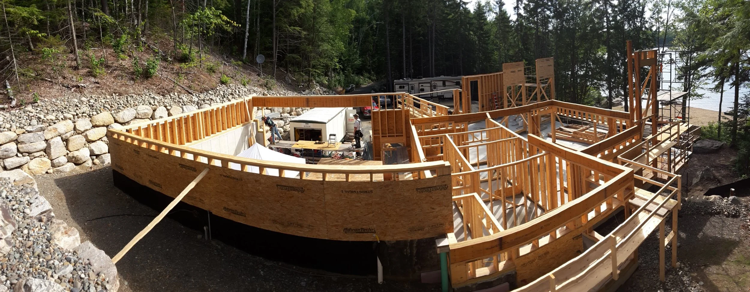 Construction site of a wooden building frame near a forest, with workers and construction materials visible.