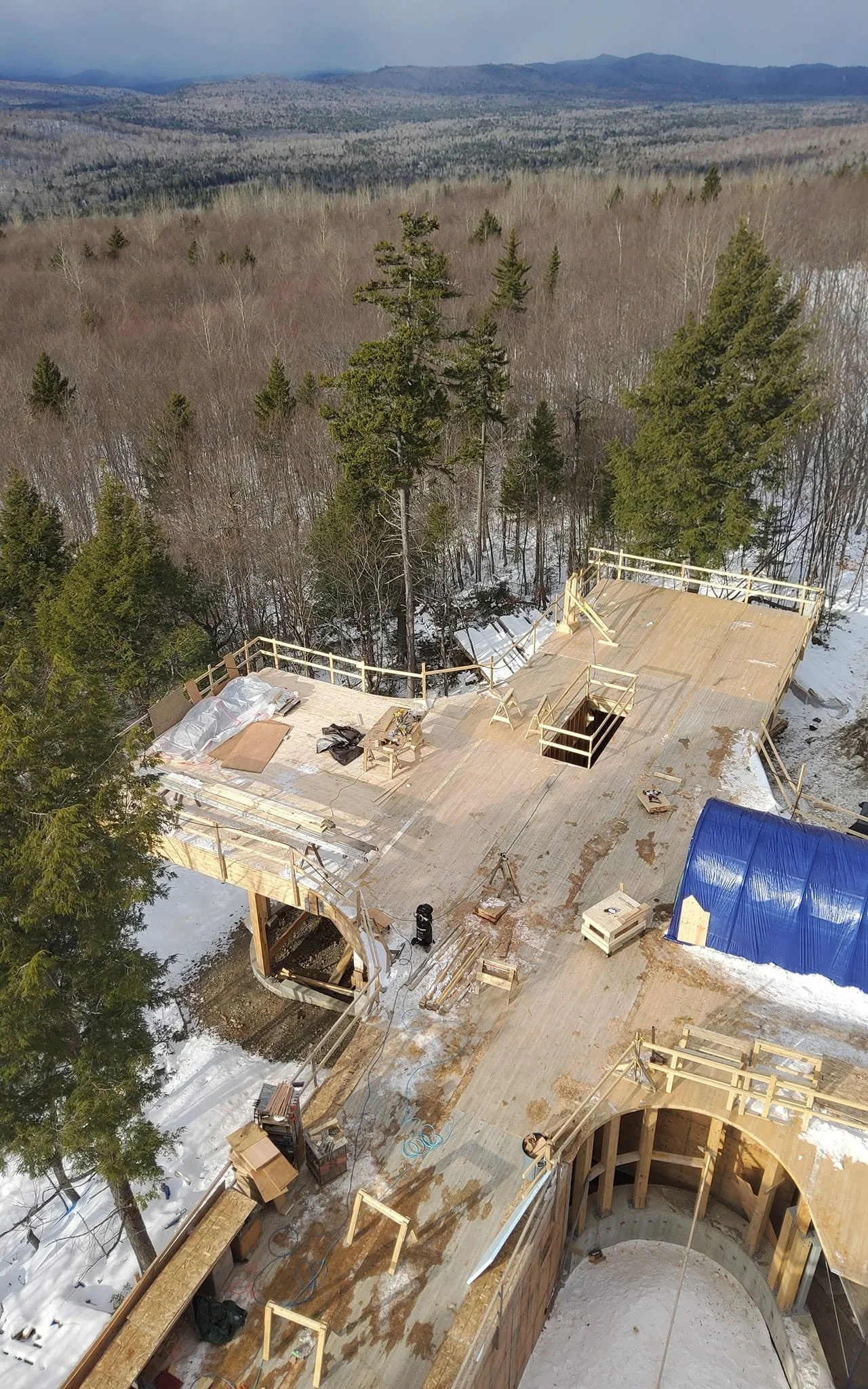 Aerial view of a building under construction in a snowy forested area, with trees, mountains in the distance, and construction materials on the site.