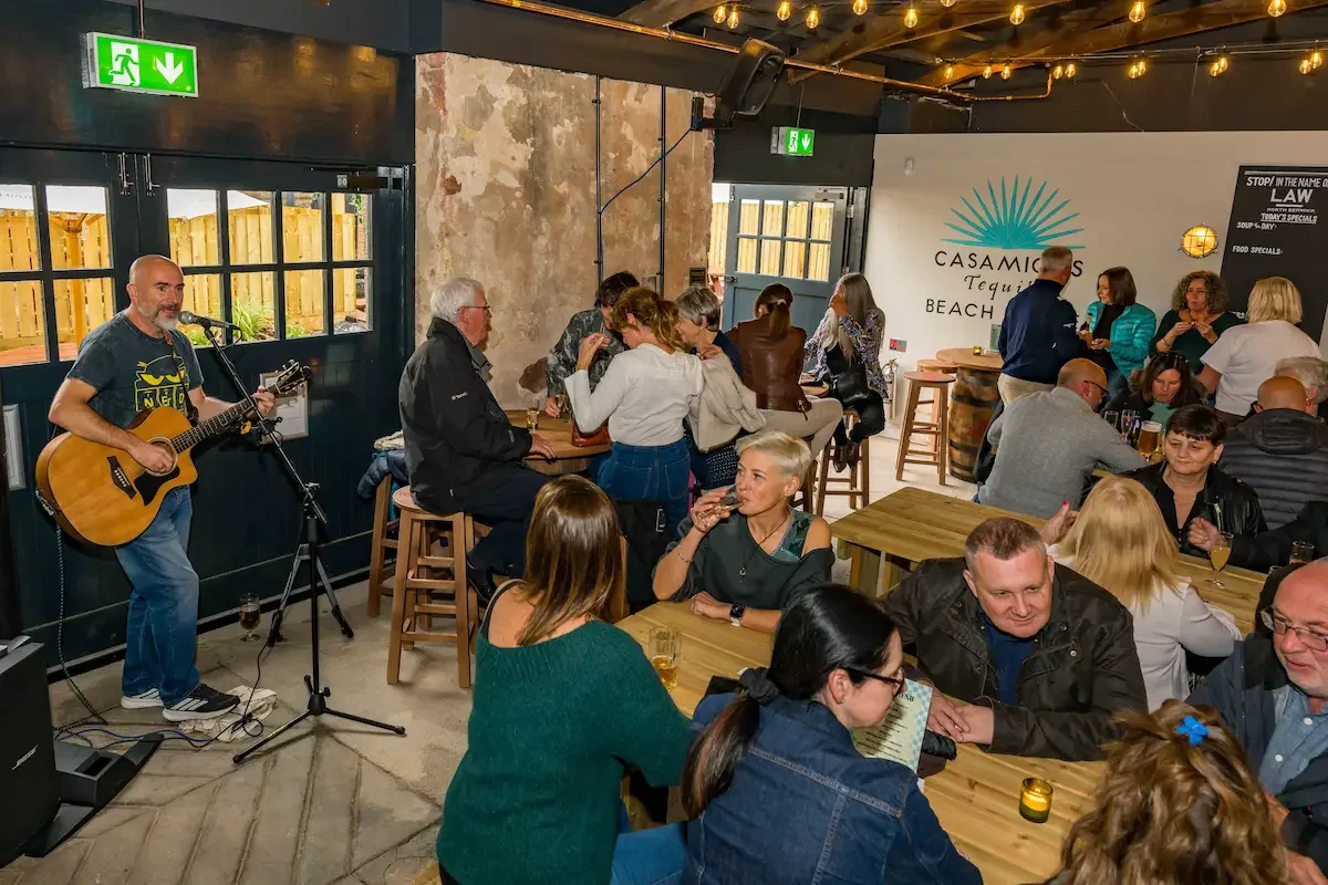 People gathered in a bar or restaurant enjoying music from a guitarist playing an acoustic guitar, with some seated at tables and others standing and chatting.