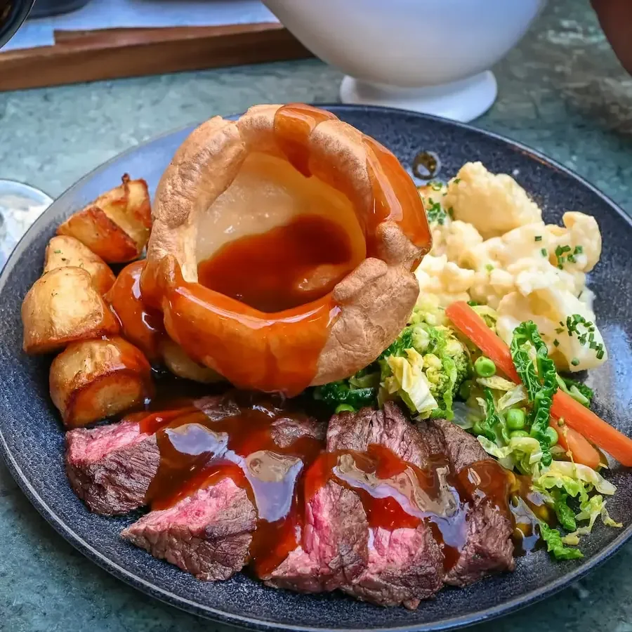 A plate of roast beef with gravy, mashed potatoes with chives, roasted potatoes, a Yorkshire pudding filled with gravy, and mixed vegetables, served at a restaurant.