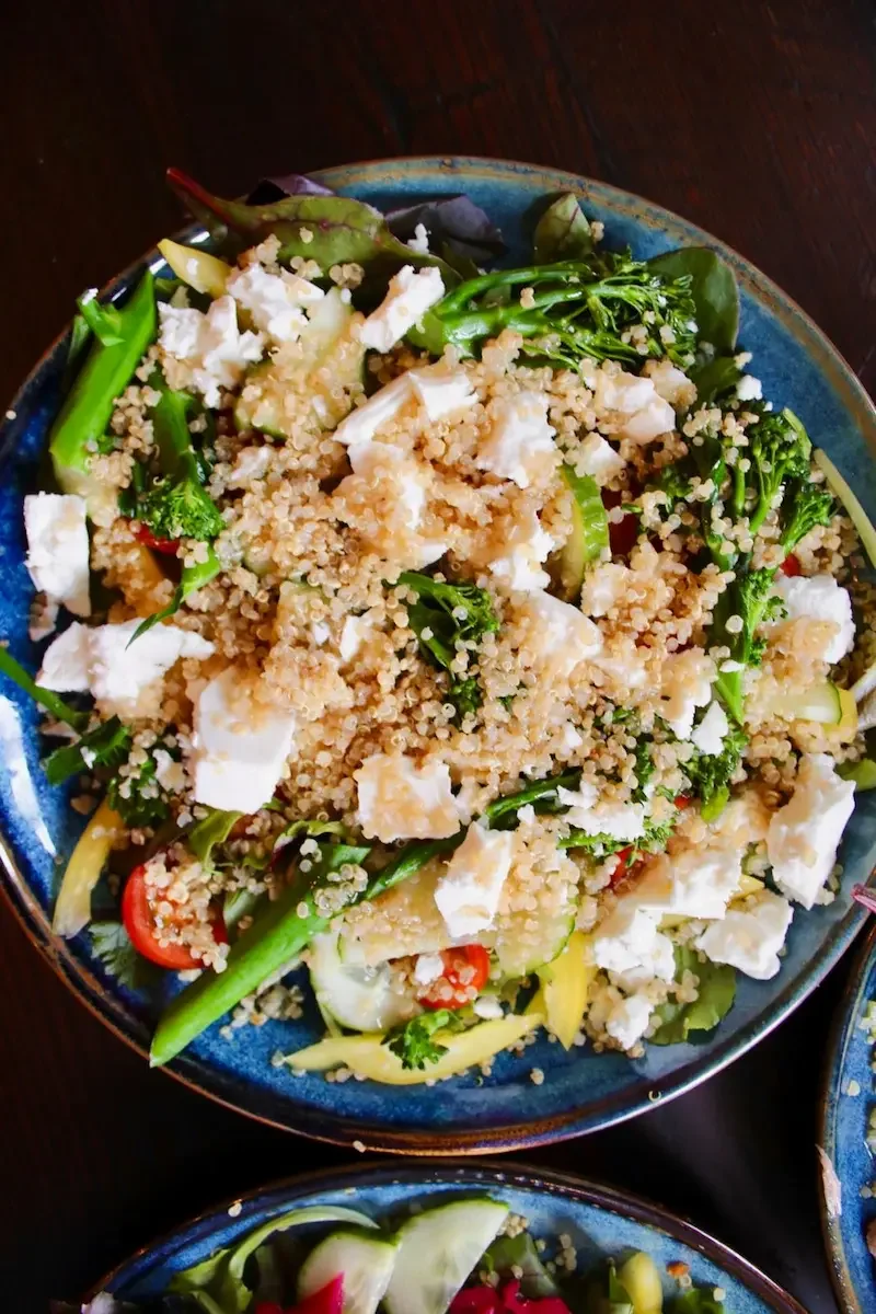 A bowl of salad with quinoa, mixed greens, cherry tomatoes, sliced cucumbers, and crumbled cheese.