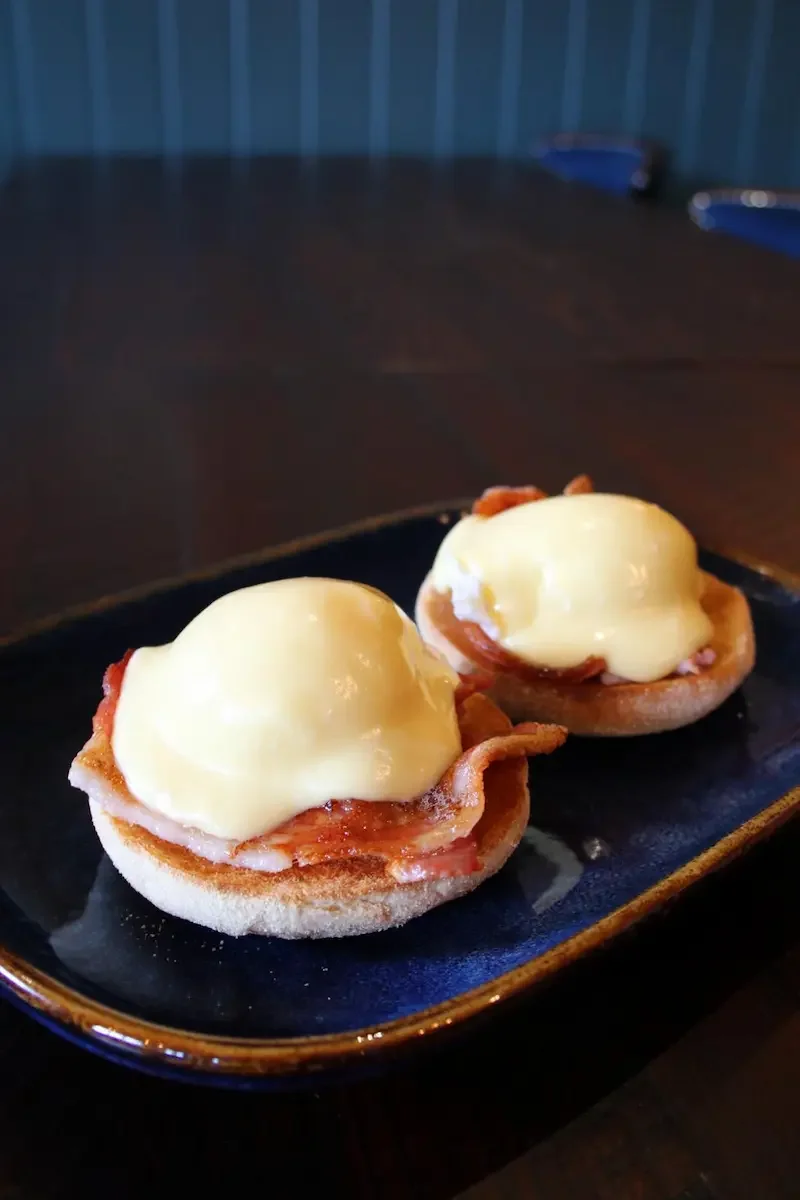 Two eggs Benedict on toasted English muffins with bacon, topped with hollandaise sauce, served on a black rectangular plate.