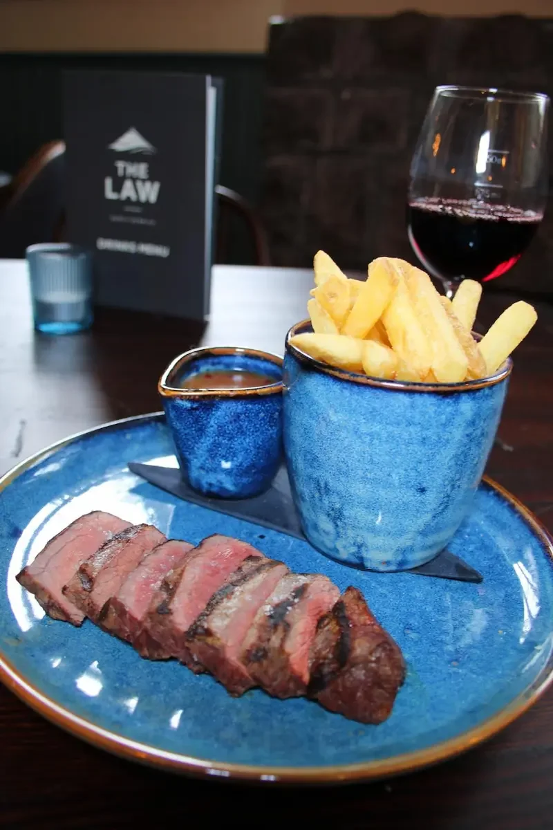 Sliced medium-rare steak with French fries in a blue ceramic cup, a small blue ceramic bowl, and a glass of red wine on a restaurant table.