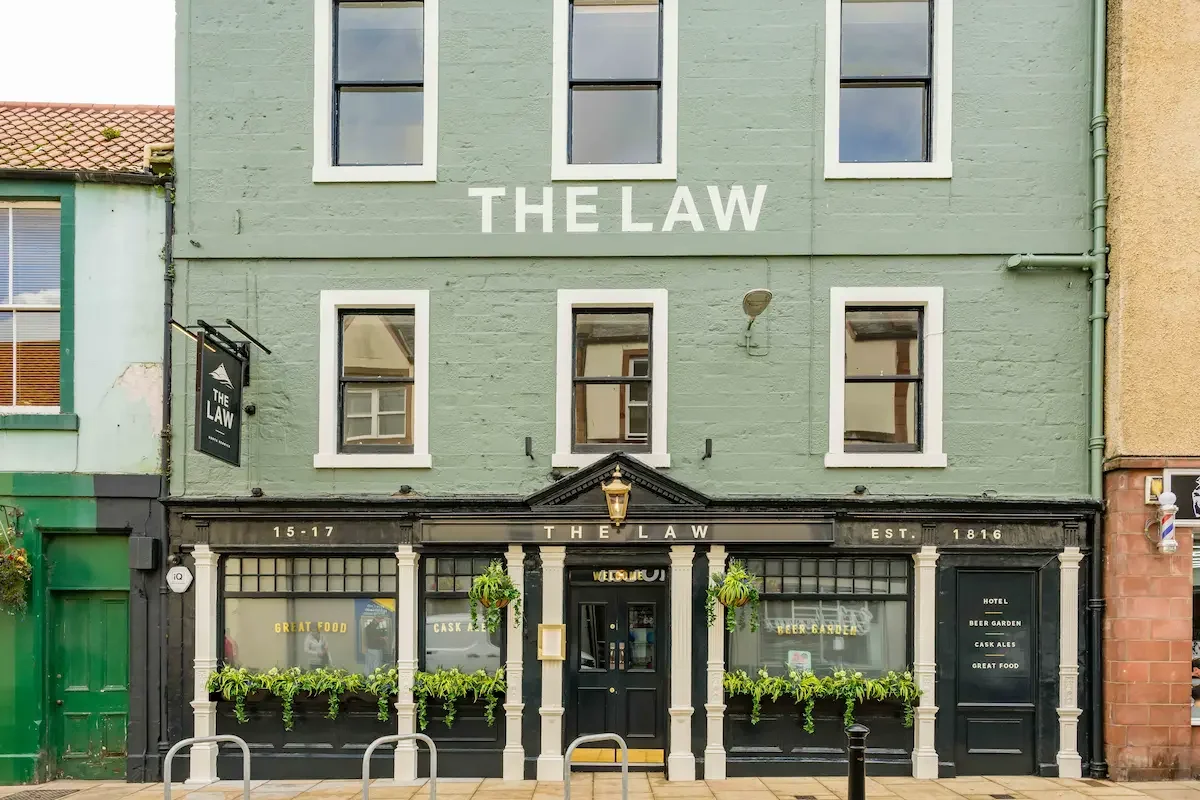 Front view of a three-story building painted in light green, with the words "THE LAW" written on the upper part of the building. The ground floor features large windows with plants and signs reading "GREAT FOOD," "CASK," "BEER GARDEN," and "HOTEL CASK ALES." The entrance is centered with a black door and the address 15-17. A small sign on the left side also says "THE LAW."