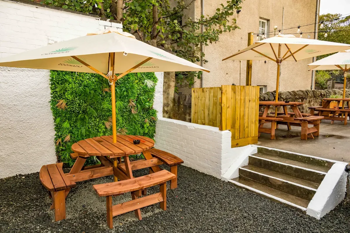Outdoor patio area with wooden picnic tables under large umbrellas, a green wall on the left, and a small set of steps leading up to another section with more picnic tables, surrounded by a white brick wall and a wooden fence.