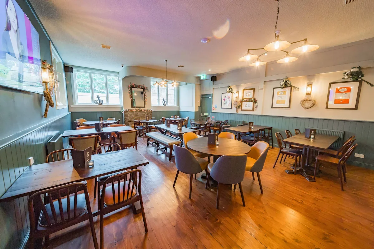 A cozy restaurant or café interior with wooden tables and chairs, decorated with framed artwork, wall-mounted lights, and flower arrangements. Large windows allow natural light to fill the space.