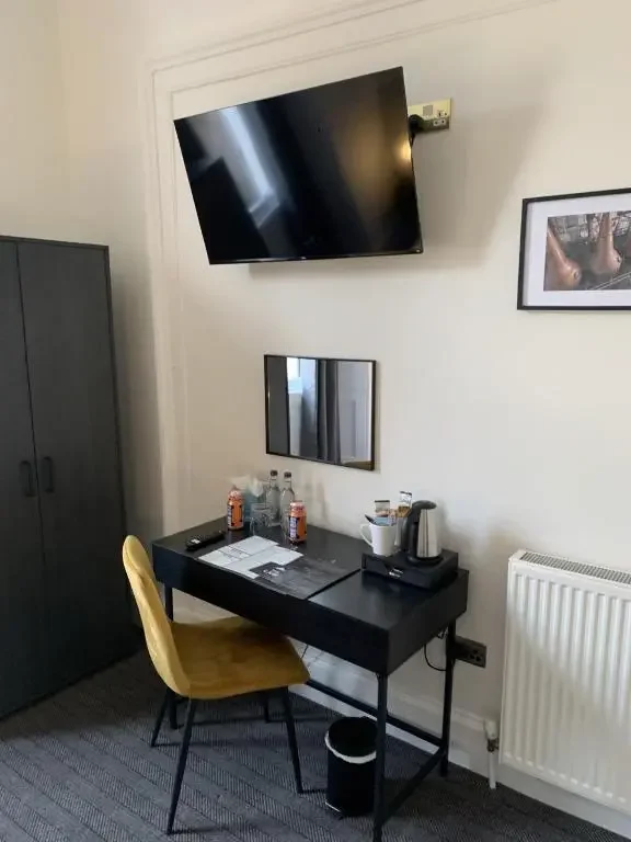 Hotel room desk with yellow chair, mirror, water bottles, glasses, electric kettle, and cups, against a white wall with a mounted TV, framed photo, and radiator.