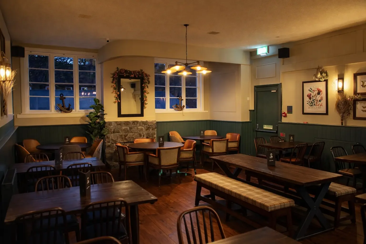 Interior of a cozy restaurant with wooden tables and chairs, warm lighting, framed art on walls, windows with a view of trees outside, and nautical decorations.