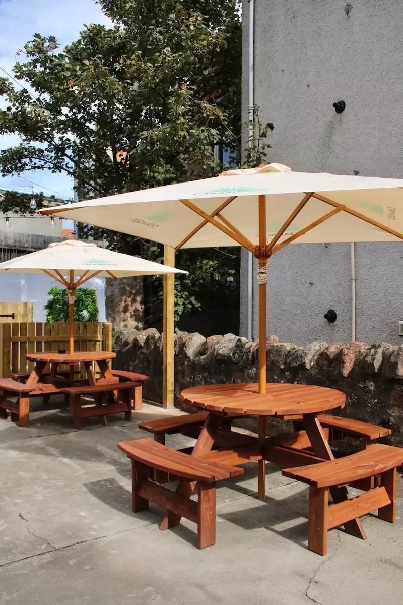 Outdoor patio with wooden picnic tables and large white umbrellas on a concrete surface, surrounded by a stone wall and a building.