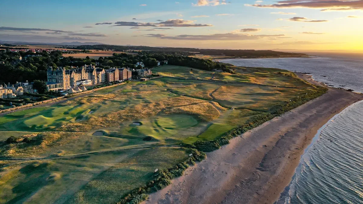 Aerial view of a coastline with a golf course adjacent to a beach, with residential buildings nearby, during sunset.