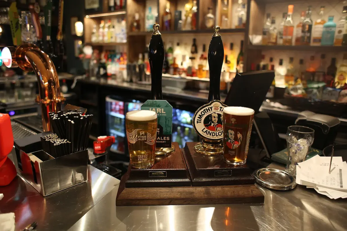 Two glasses of beer on a bar counter, each with a tap handle behind it, with bar supplies and beyond in the background.