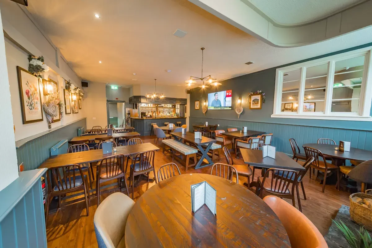 Empty cozy restaurant with wooden tables, chairs, wall art, and a bar in the background.