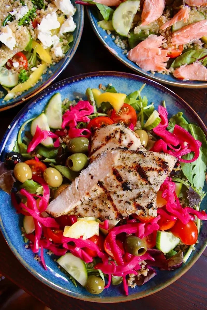 A colorful salad with grilled chicken slices, cherry tomatoes, cucumbers, olives, pickled vegetables, and mixed greens in a blue bowl.