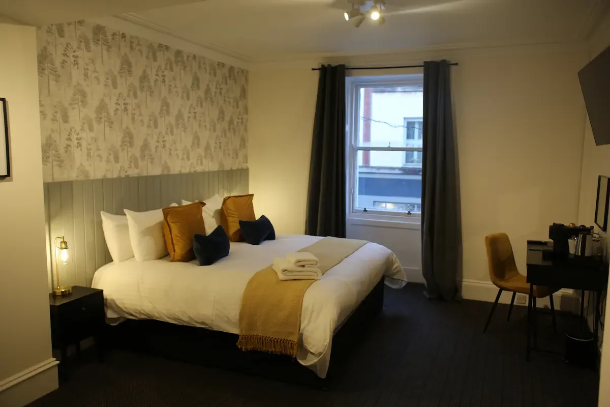 Hotel room with a large bed, decorative pillows, and a window with curtains, a small desk with a chair, and towels on the bed.