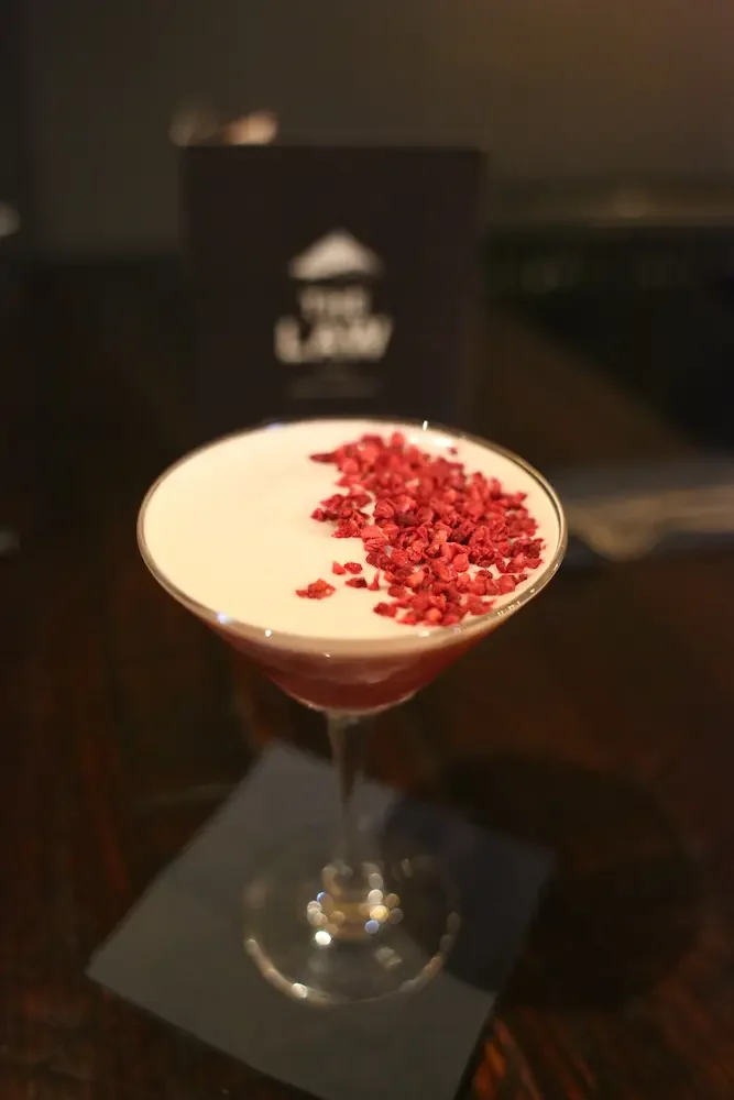 A cocktail in a martini glass with a creamy white top layer and red crushed topping, sitting on a black napkin on a wooden table.