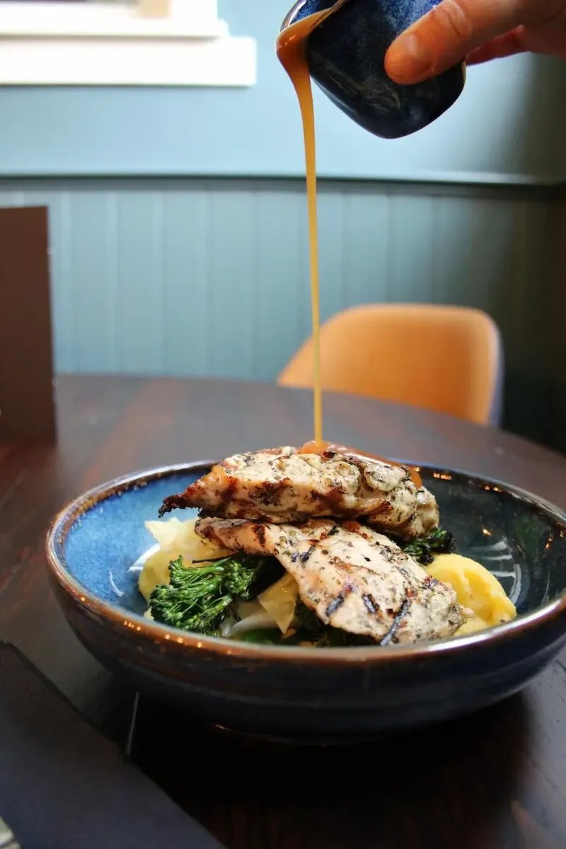 Grilled chicken pieces with vegetables on a plate, with gravy being poured over them from a cup.