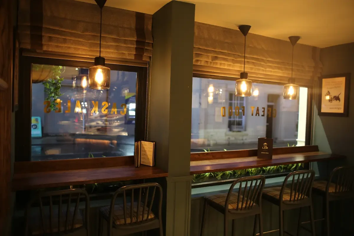 Interior of a cafe with two windows, wooden counter seating with four chairs, hanging pendant lights, and framed art on the wall.