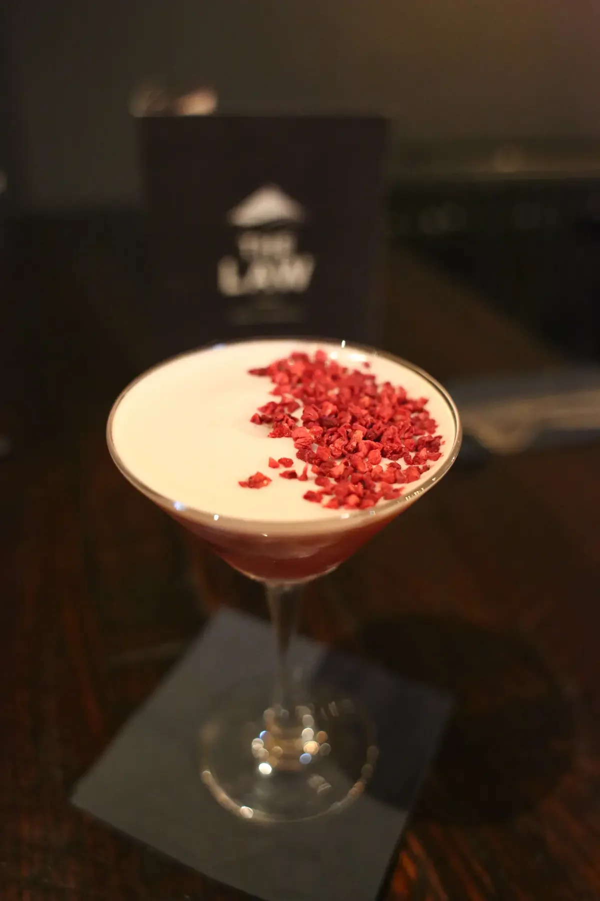 A cocktail glass with a layered drink topped with red garnish, placed on a black napkin on a wooden table in a dimly lit room.