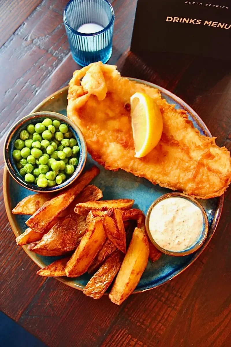 A plate of fried fish with lemon, potato wedges, green peas, tartar sauce, and a glass of milk on a wooden table.