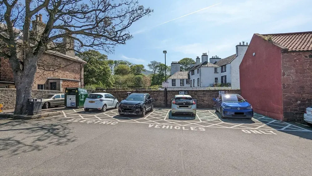 A small parking lot with five cars, an electric vehicle charging station, a large tree, a brick wall, and residential buildings in the background.