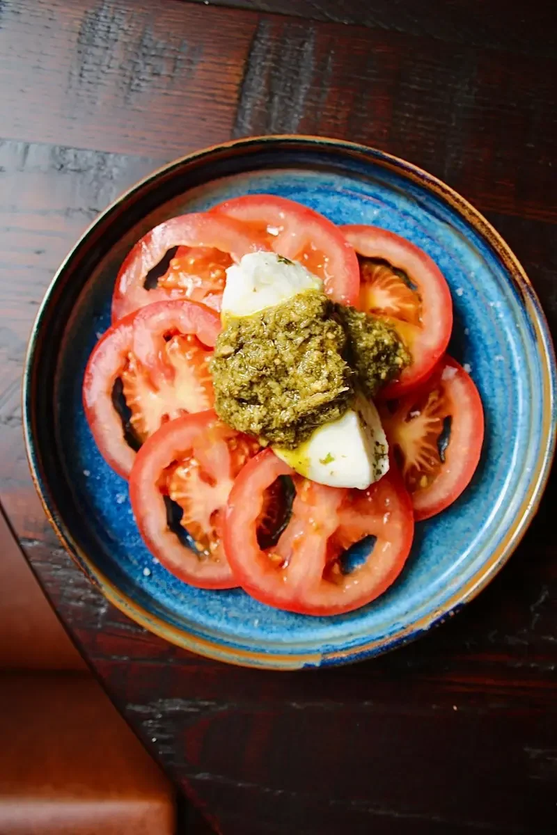 A blue ceramic bowl containing sliced tomatoes, hard-boiled eggs, and a dollop of greenish pesto.