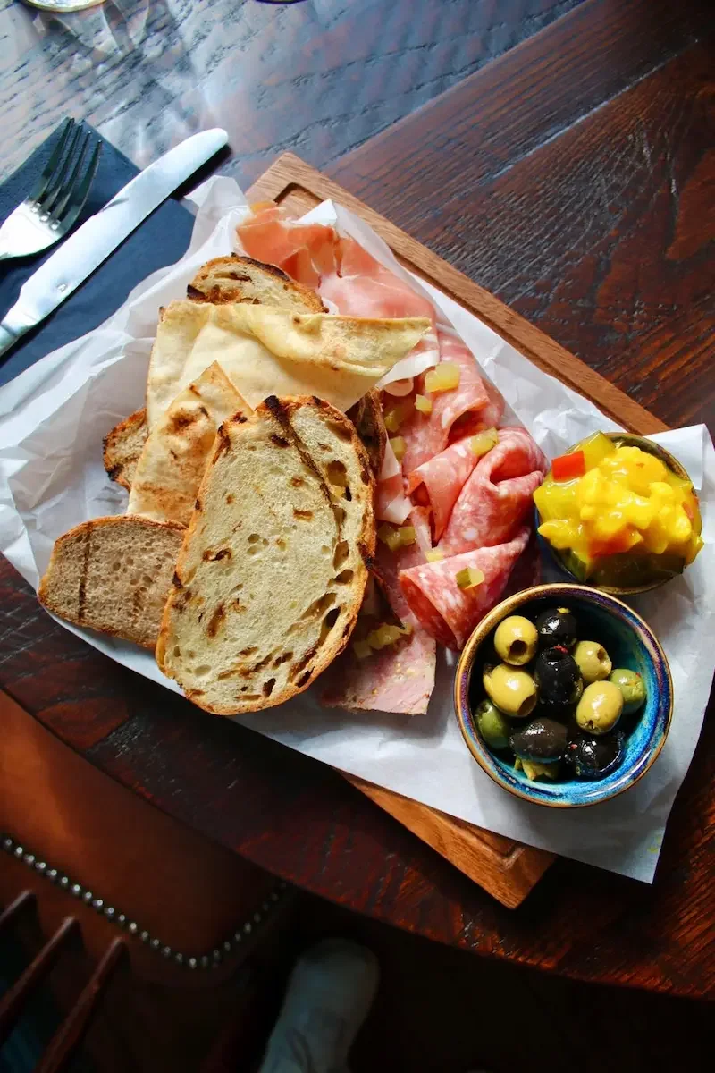 A wooden tray with a variety of bread slices, cured meats, and small bowls of olives, pineapple, and diced tomatoes with peppers.