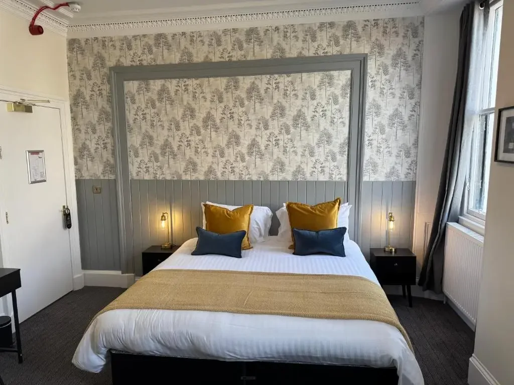A neatly made bed with white linens, four pillows (two gold and two dark blue), two bedside tables with small lamps, a window with curtains, and a framed picture on the wall inside a hotel room.