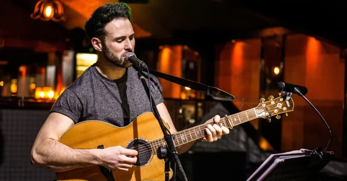 A man playing acoustic guitar and singing into a microphone in a dimly lit indoor setting.