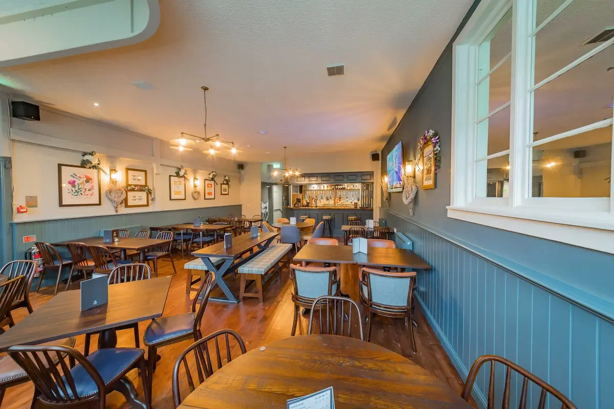 Interior of a cozy restaurant with wooden tables and chairs, decorated with framed artwork and flowers, bar area in background, warm lighting, and blue paneled walls.