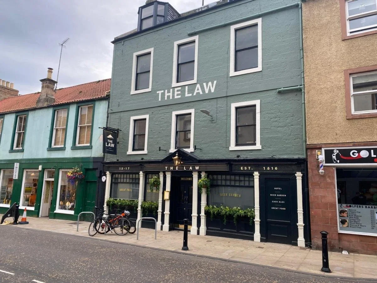 A three-story building painted in light green with white window trims, housing a pub called "The Law" established in 1816. The pub has black storefront accents, hanging flower baskets, and a sign. Bicycles are parked in front on a city sidewalk. To the right, part of another building and a barbershop sign are visible.