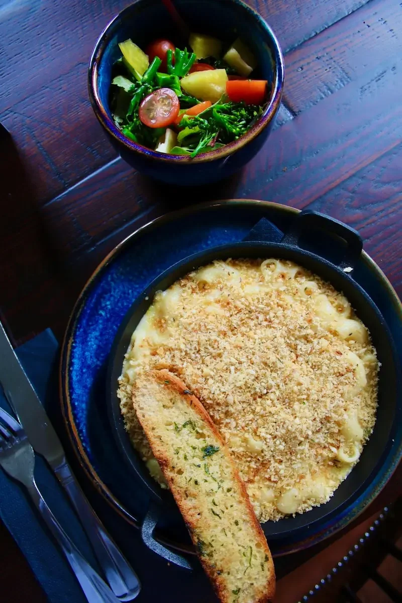A bowl of baked macaroni and cheese topped with breadcrumbs and a slice of garlic bread, accompanied by a small bowl of mixed salad with cherry tomatoes, cucumber, broccoli, and carrots on a wooden table.
