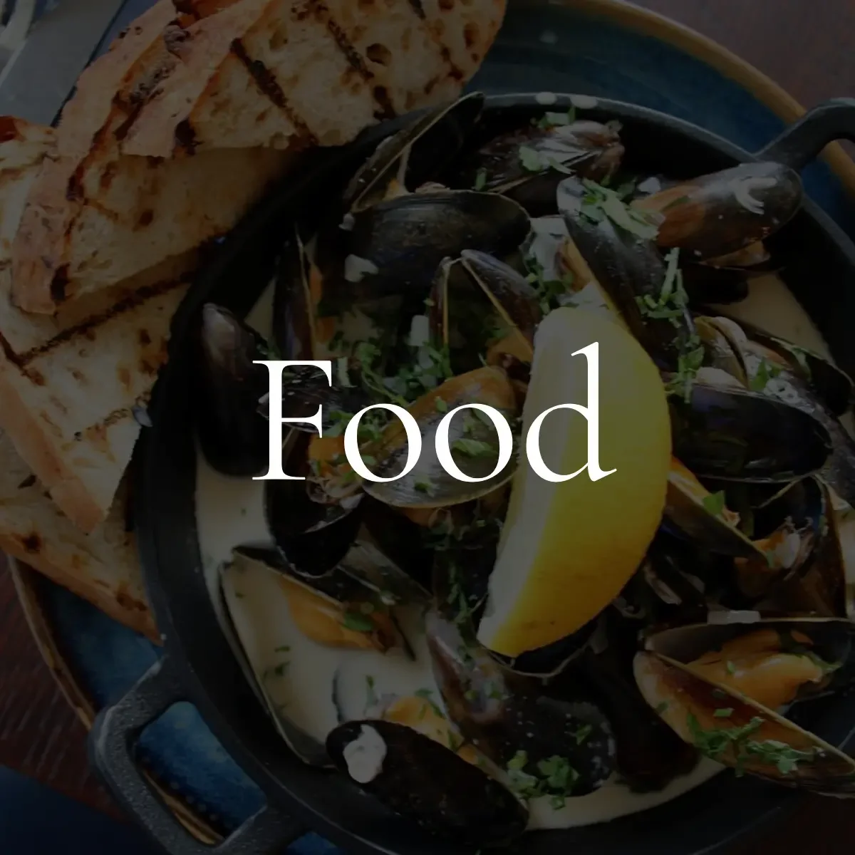 A dish of cooked mussels garnished with chopped herbs and a lemon wedge, served with toasted bread slices.