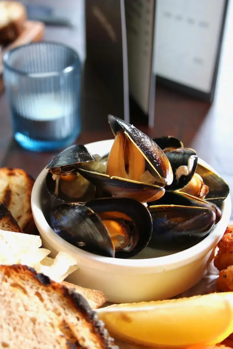 A bowl of steamed mussels with lemon wedges on a plate with grilled bread and fried food, on a table with a glass of water and a blurred background.