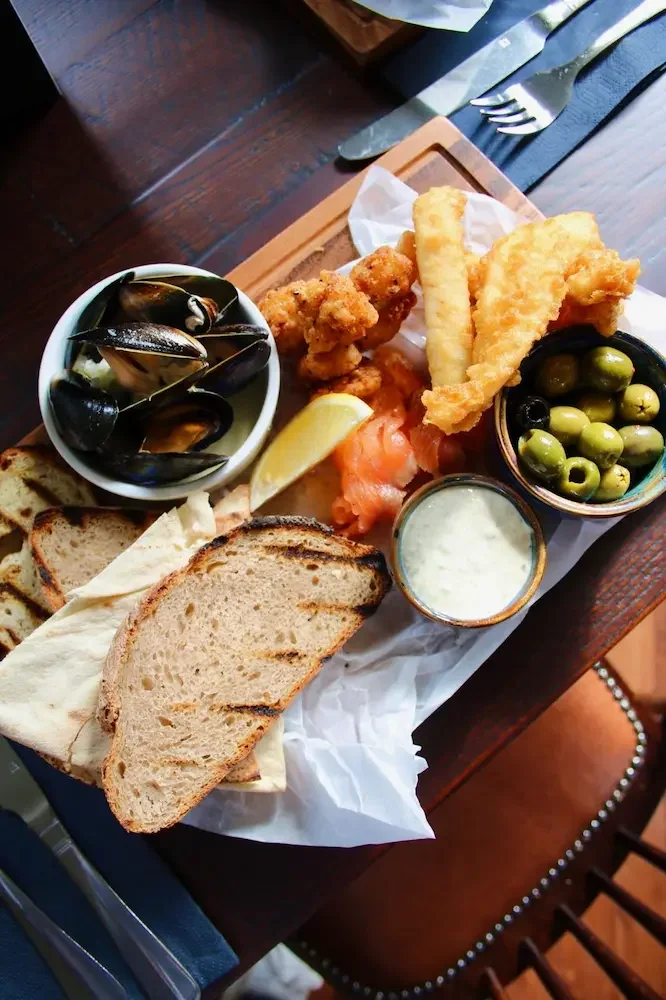 A serving of fried fish fillets, French fries, a bowl of green olives, a bowl of mussels, slices of bread, lemon wedge, and tartar sauce on a wooden table.