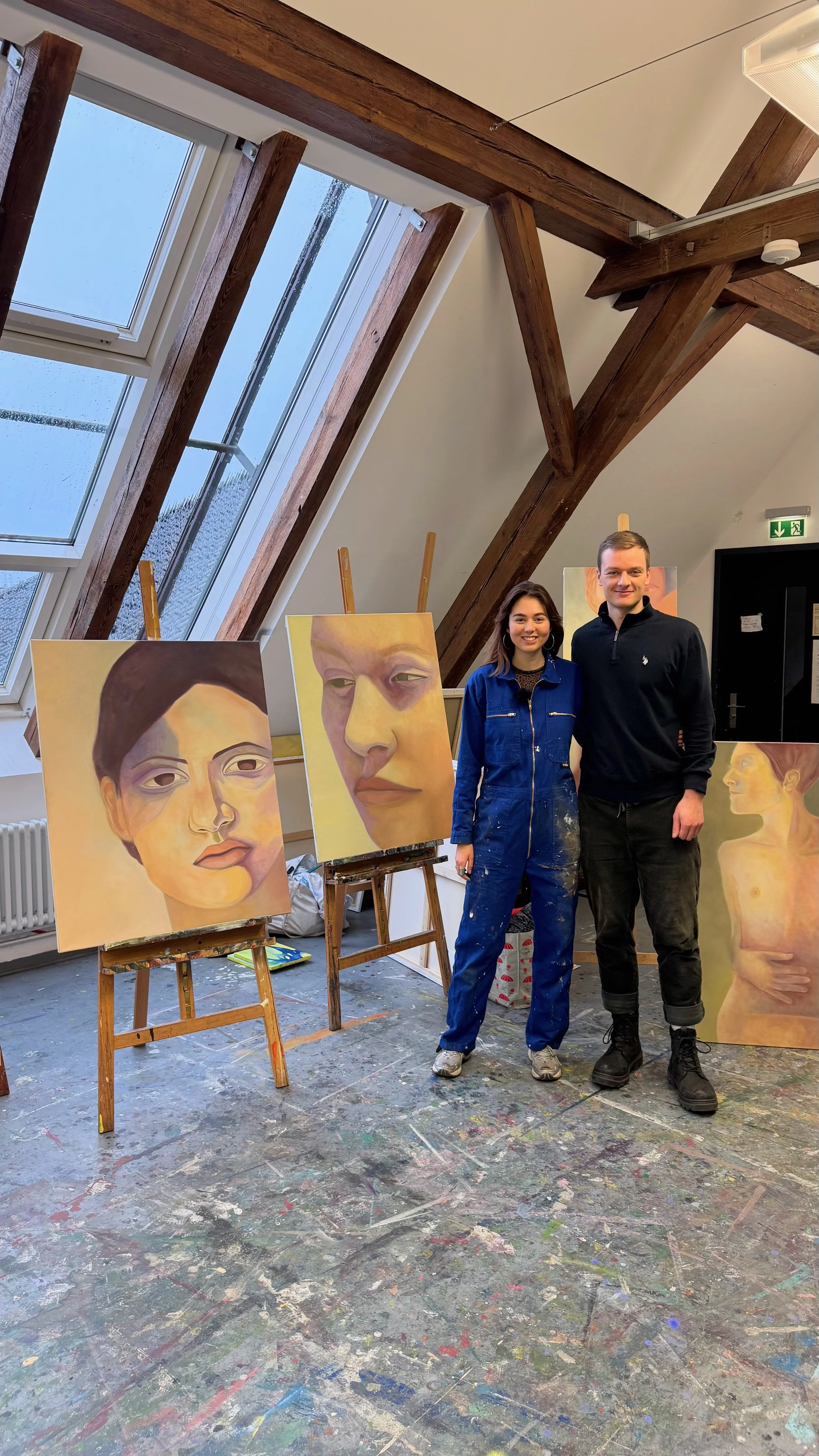 Zwei Künstler stehen in einem Atelier, umgeben von ihren Gemälden. Die Gemälde sind Porträts in einem abstrakten, impressionistischen Stil. Das Atelier hat große Dachfenster, die verteilt natürliches Licht in den Raum lassen, und der Boden ist mit Farben und Leinölfarben bespritzt.