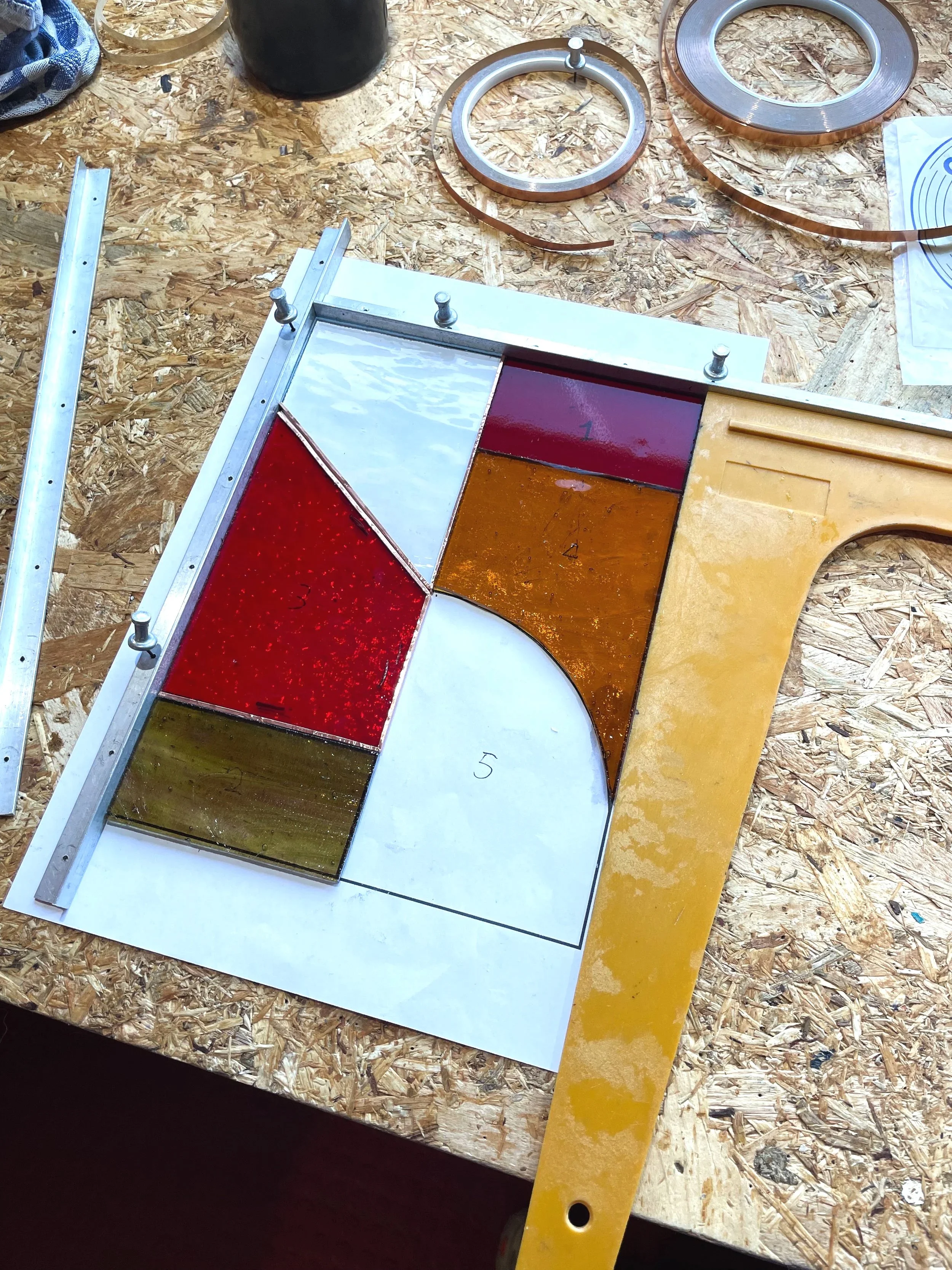 Een werkbank met verschillende gereedschappen en glas in lood materialen, waaronder gekleurde lamellen en metalen ringen, op een houten werkblad.