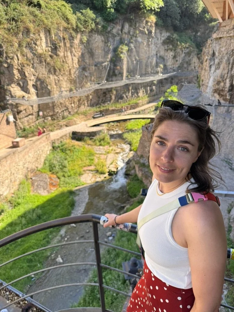 De glazenier Een jonge vrouw met een witte top en een rode polkadotrok staat op een uitkijkpunt met een ijzeren balustrade, met een bergachtig landschap en een rivier eronder.